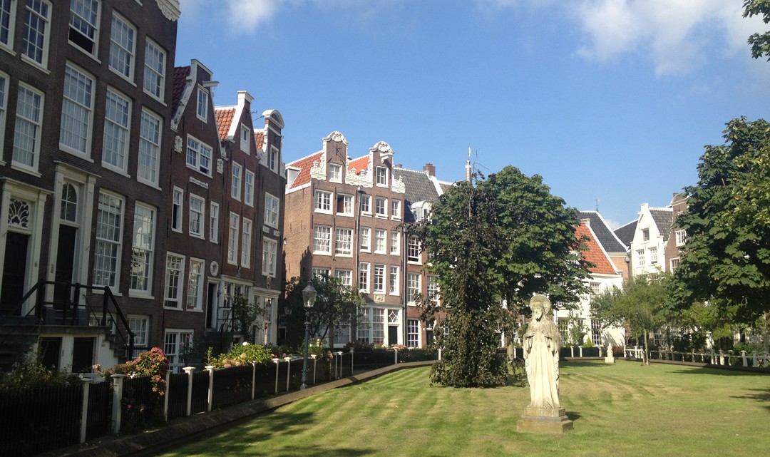 Green grass surrounded with typical Amsterdam gable houses. Trees and a statue sit in the middle of the garden