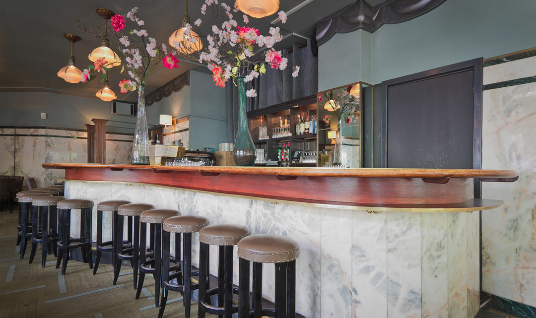 A row of barstools along a curvy bar and high vases with flowers on the light wooden bench top