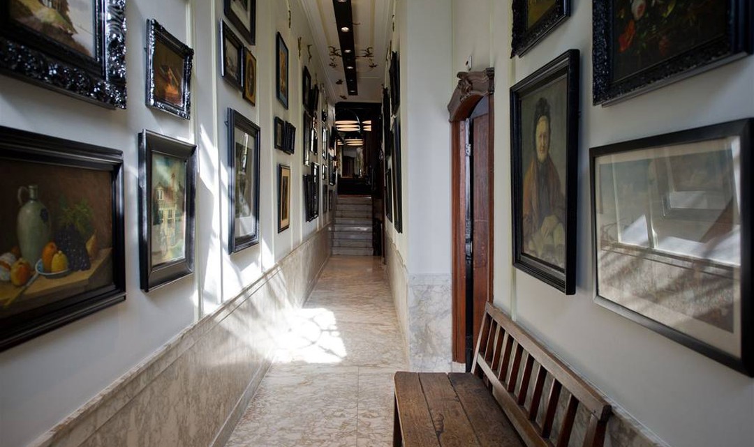 A long, narrow hallway with marble floors. The wall filled with historic portraits, still lifes and other paintings