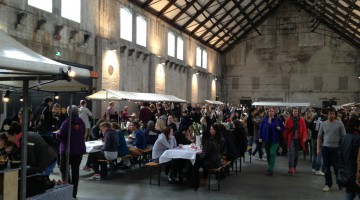 Food stands and long table with snacking people in an enormous industrial looking hall