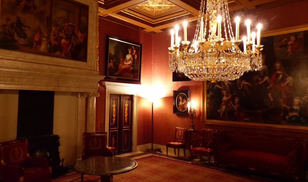 A richly decorated red-brown room with one chandelier and walls full of paintings