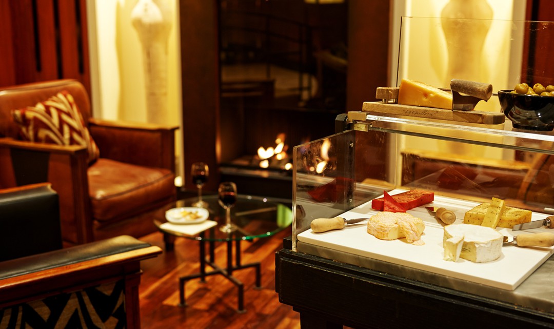 A cheese trolley with various cheeses; in the background two leather chairs in front of an open