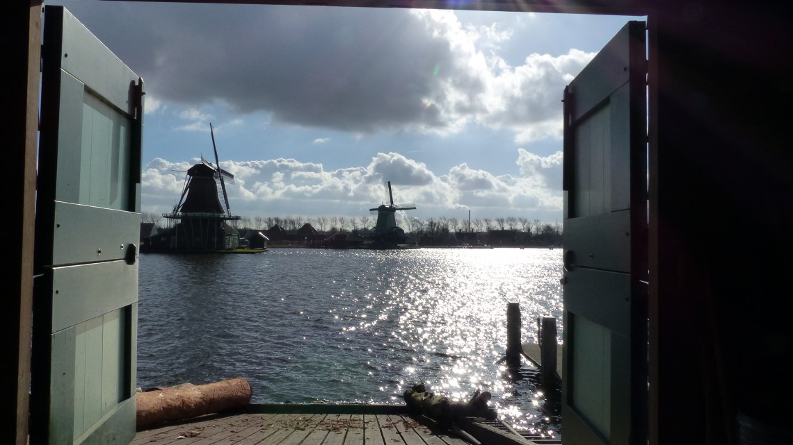 A view from open doors to two windmills along the water with Dutch clouds and sun