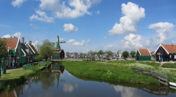 A view over the water with a blue Dutch sky with friendly clouds, a windmill in the background and typical Zaanse houses left and right